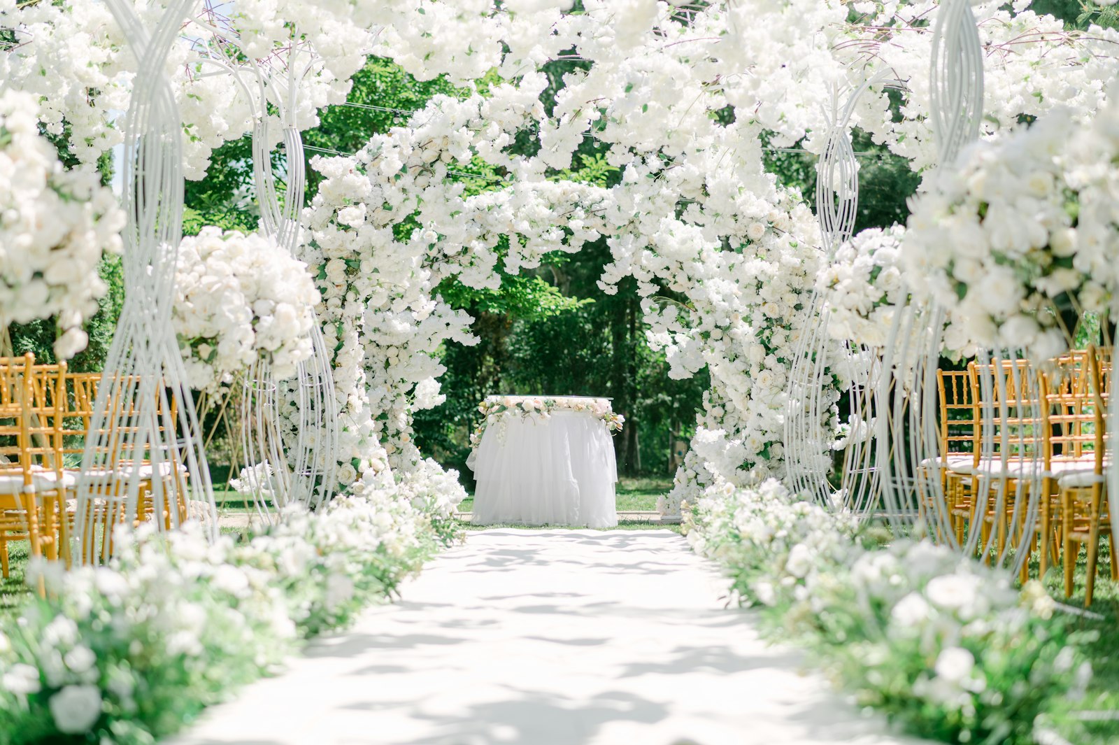 Gartenhochzeit mit weißem Blumenbogen über dem Gang und goldenen Stühlen